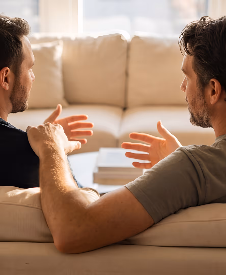 Two men sitting on a beige sofa engaged in a conversation, using hand gestures.