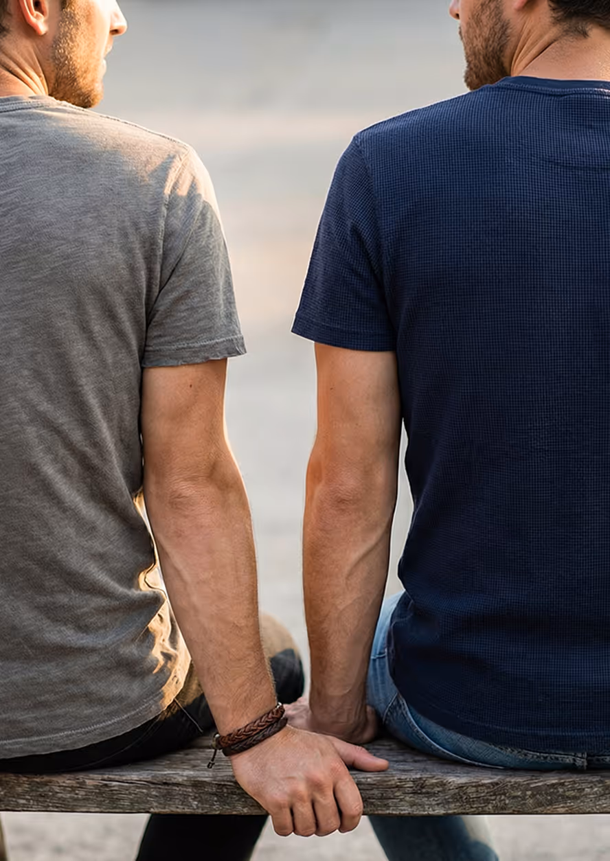 Two men sitting side by side on a wooden bench holding hands behind their backs.