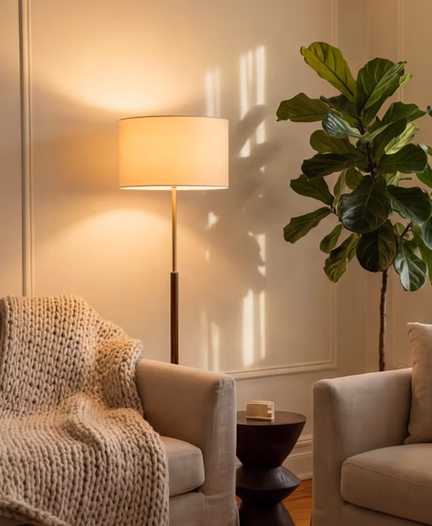 Cozy living room corner with cream armchairs, chunky knit blanket, floor lamp, small wooden side table, and a tall leafy plant.