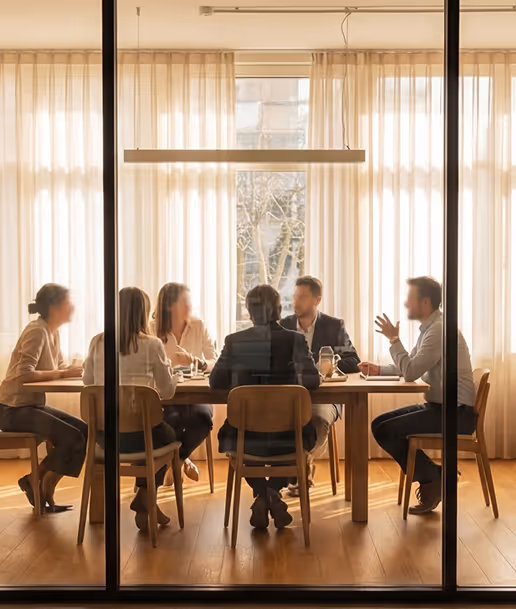 Five people having a meeting around a wooden table in a bright room with large windows and sheer curtains.