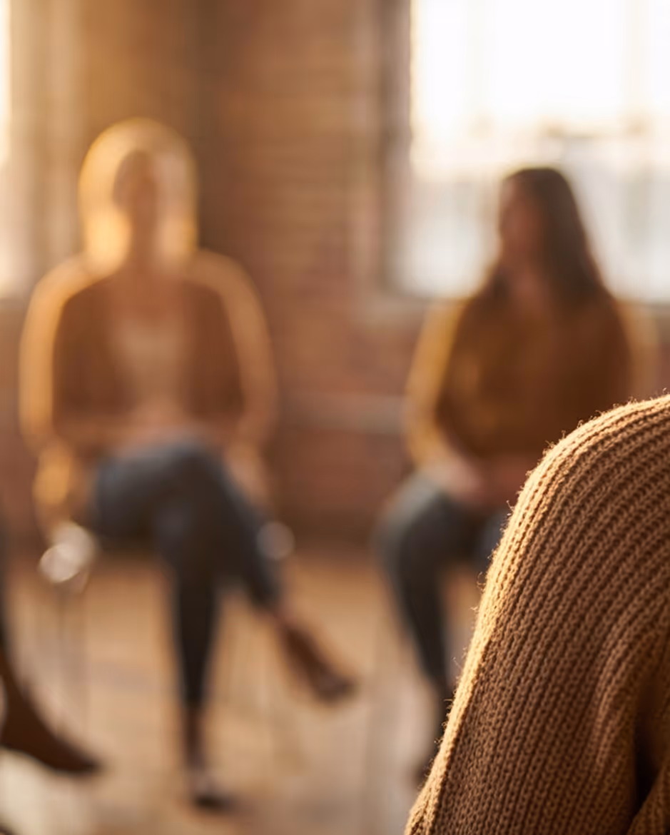 People sitting in a circle during a group therapy or support session in a warmly lit room.