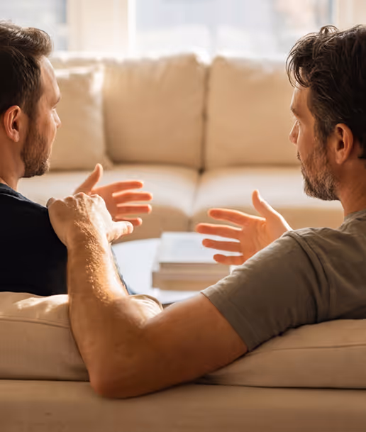 Two men sitting on a beige couch engaged in a conversation, one gesturing with hands and the other with a hand on his shoulder.
