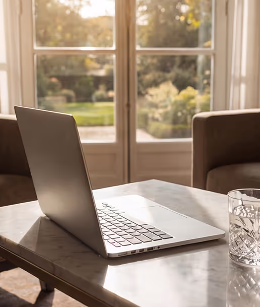 Silver laptop open on a marble table with a glass of water next to it, sunlight streaming through a large window overlooking a garden.