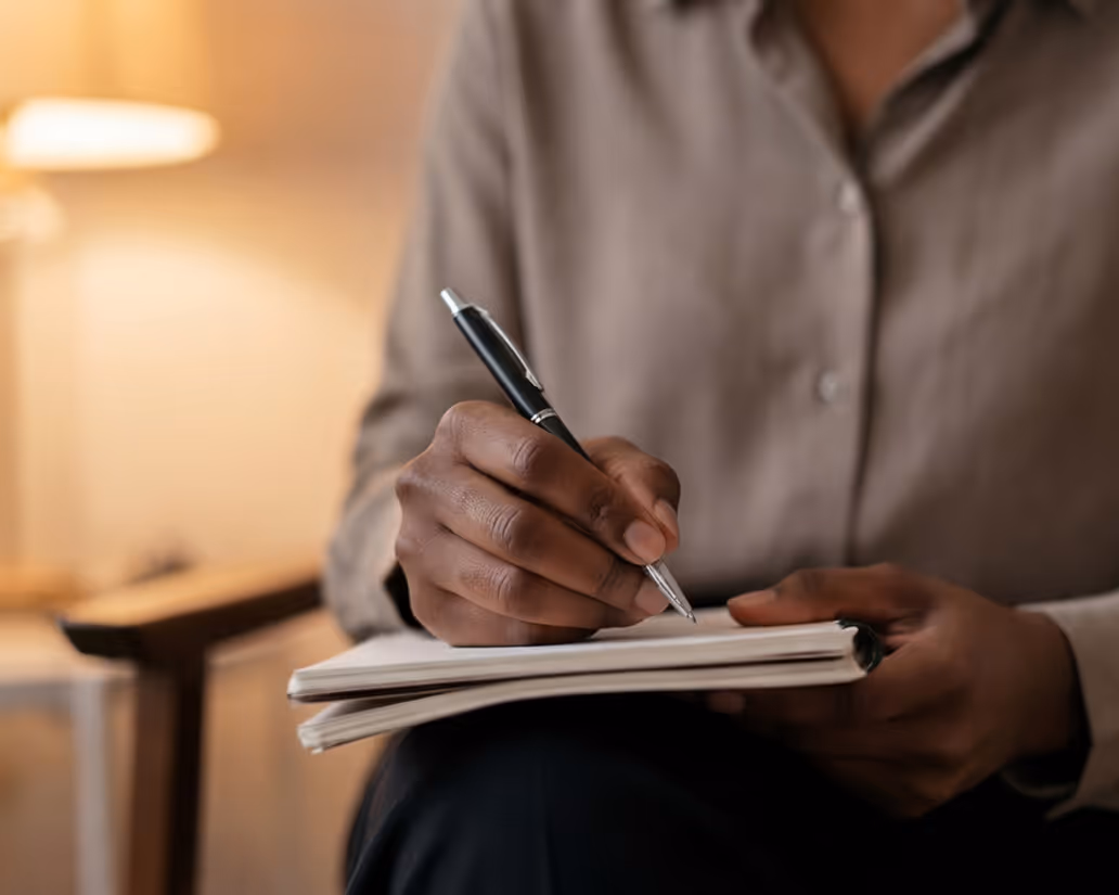 Person in beige shirt writing in a notebook with a black pen in a softly lit room.