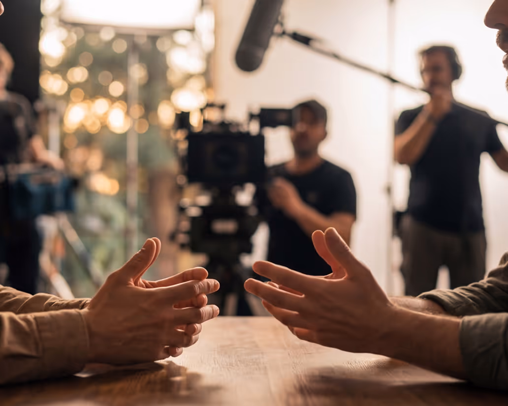 Two people gesturing with their hands across a wooden table during a filmed conversation with a camera crew and boom mic in the background.