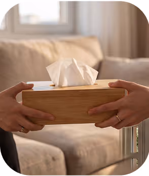 Two hands holding a wooden tissue box with tissues on a beige couch background.