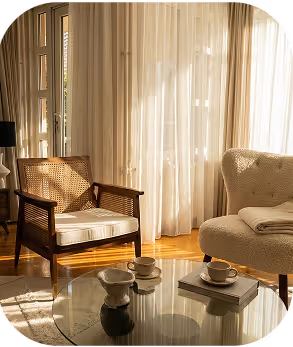 Sunlit cozy living room with a cushioned wooden chair, a soft armchair, and a glass coffee table holding teacups and a small vase.