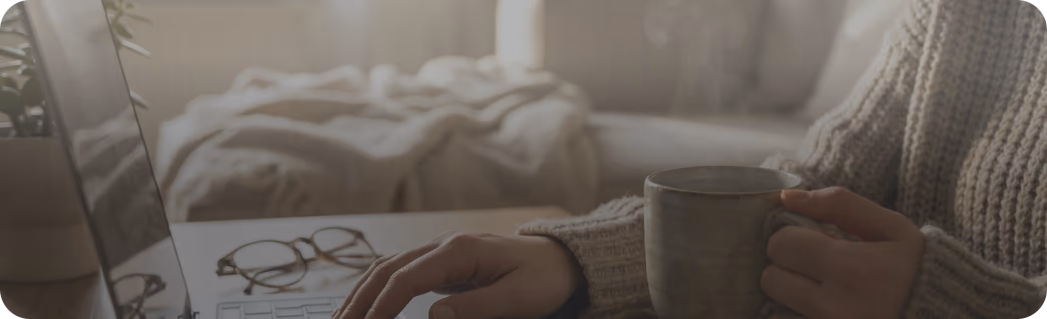 Person in a knitted sweater typing on a laptop while holding a steaming mug with glasses and a cozy blanket in the background.