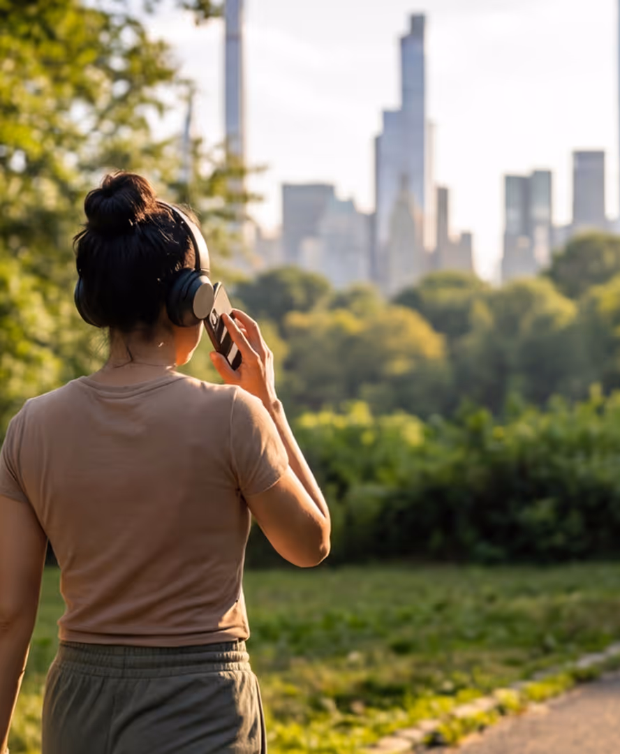 Person wearing headphones and talking on a phone in a green park with a city skyline in the background.