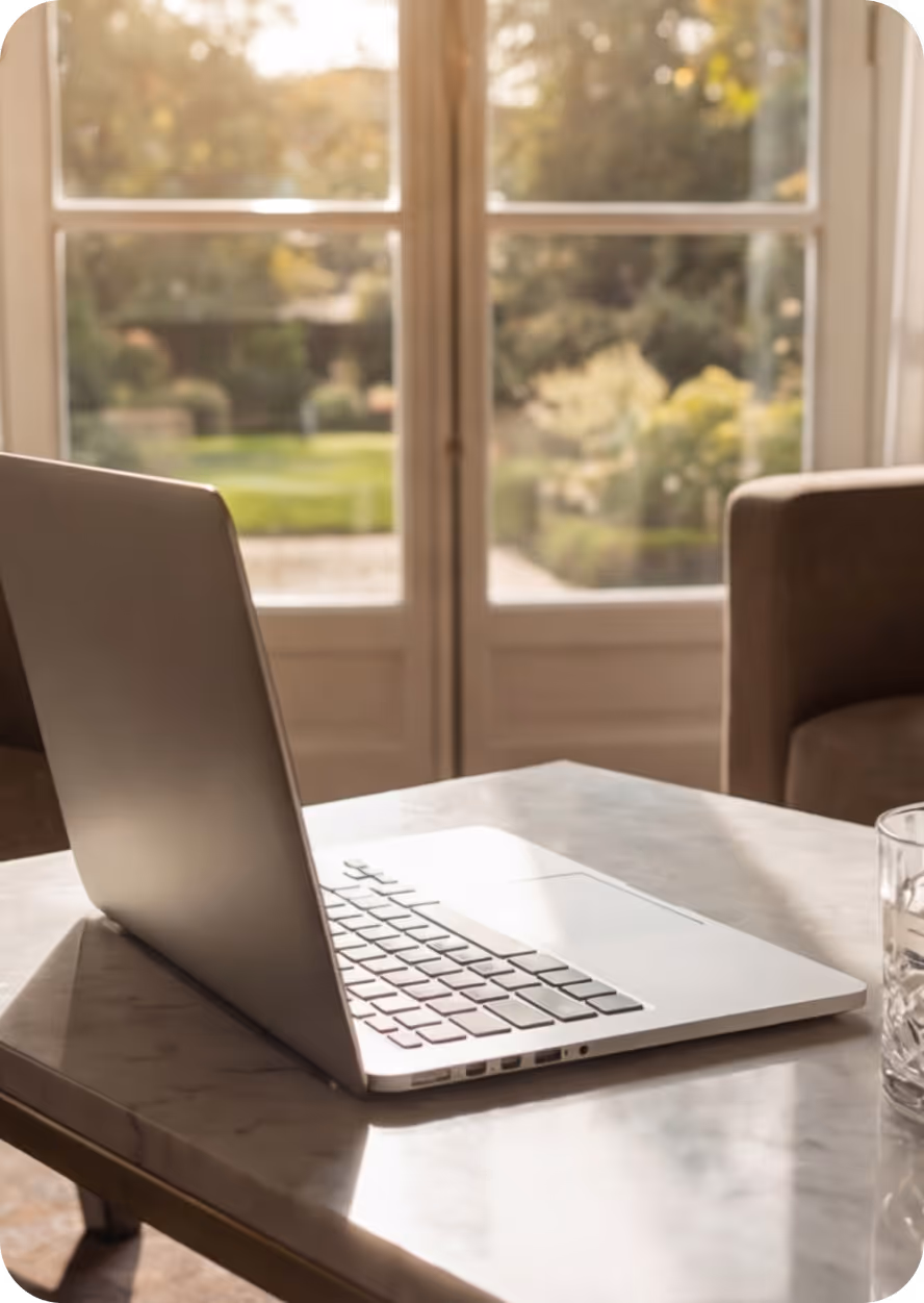 Silver laptop on a marble table near a glass of water with a sunlit garden visible through a window in the background.