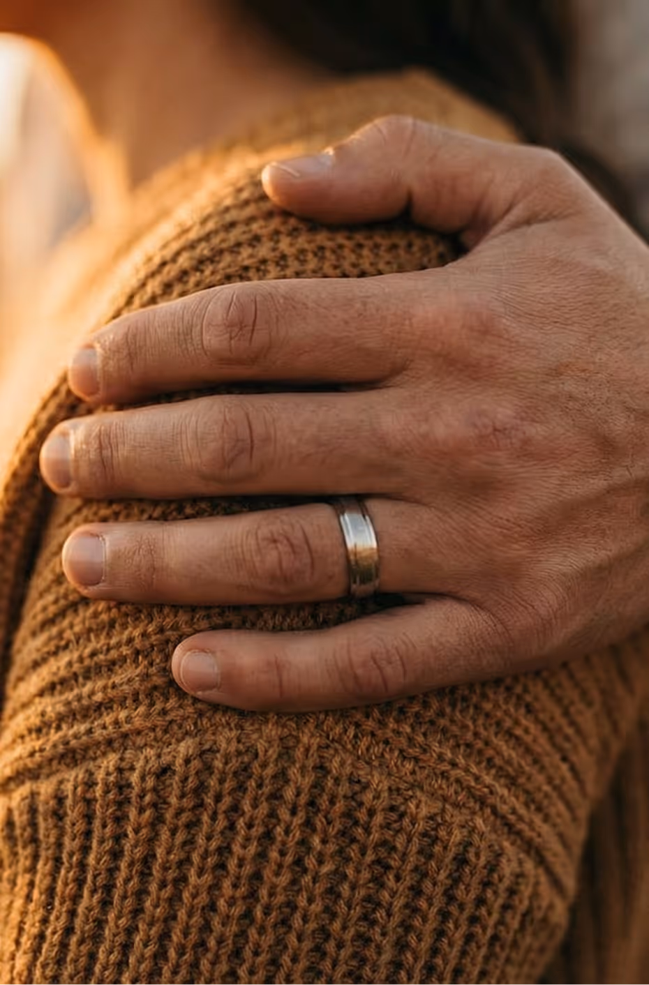 Close-up of a man's hand with a silver wedding ring resting on a shoulder wearing a brown knitted sweater.