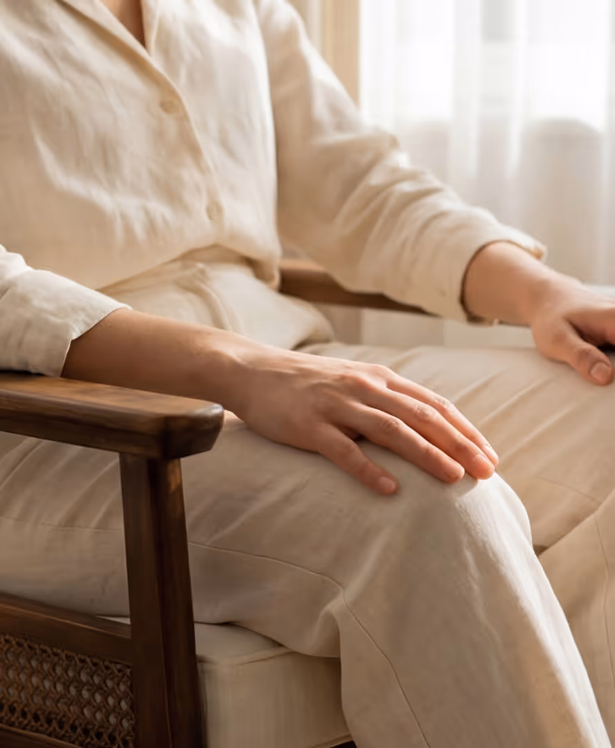 Person wearing beige linen shirt and pants sitting on a wooden chair with hand resting on knee.