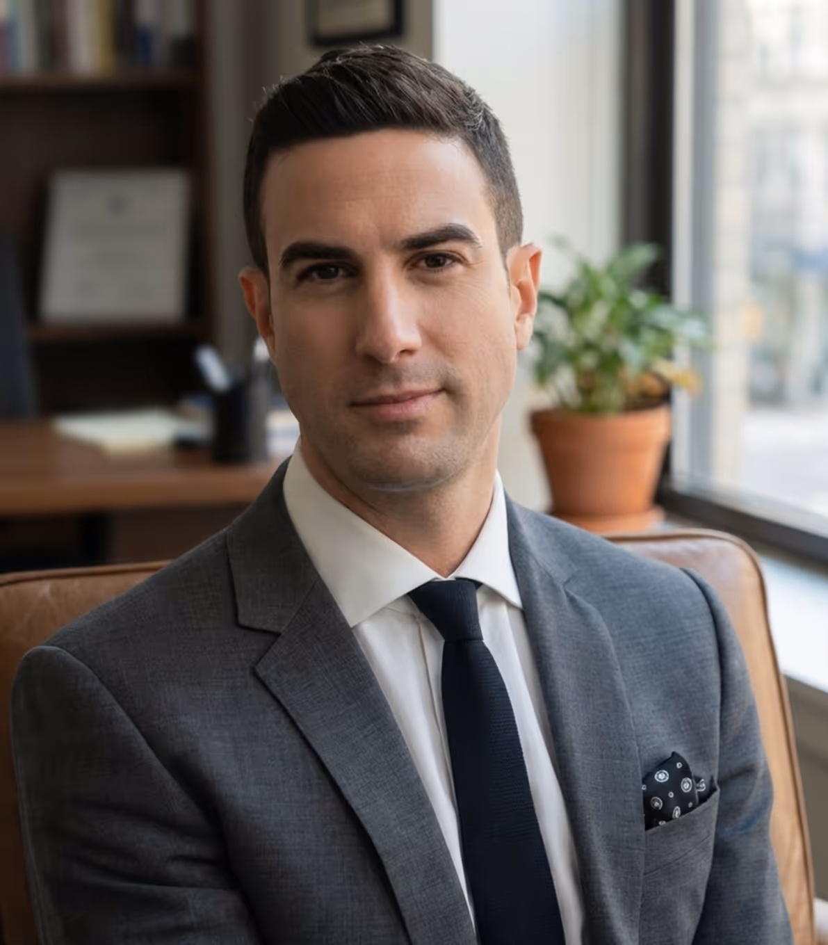 Serious man in a gray suit and black tie sitting indoors with a plant and window in the background.