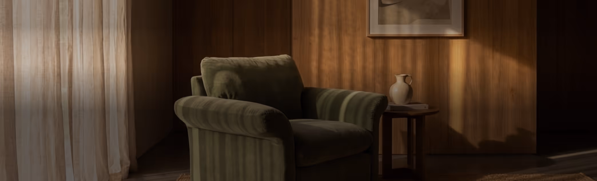 Green armchair next to a wooden side table with a white vase and books in a warmly lit room with sheer curtains.