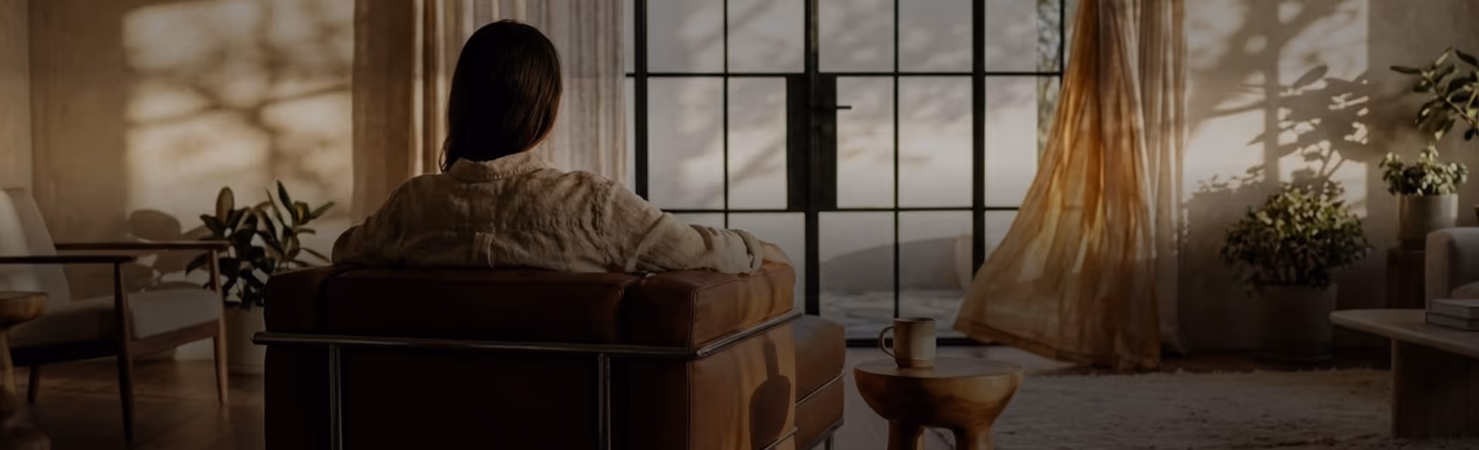 Person sitting on a brown leather chair facing large window with sunlight casting shadows of plants indoors.