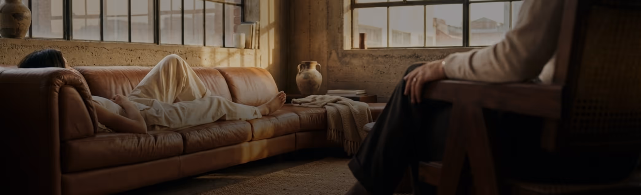 Person lying down on a brown leather couch in a warmly lit room with large windows, and another person sitting nearby.