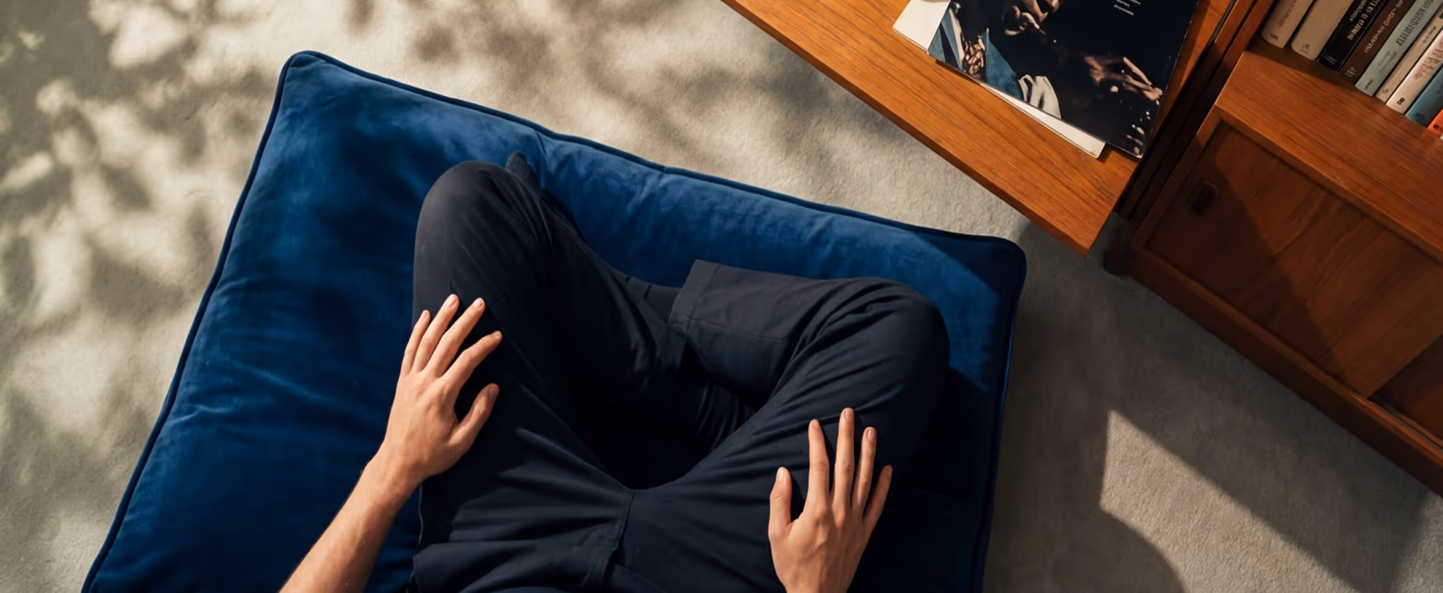 Person sitting cross-legged on a blue cushion next to a wooden bookshelf and a coffee table with books.