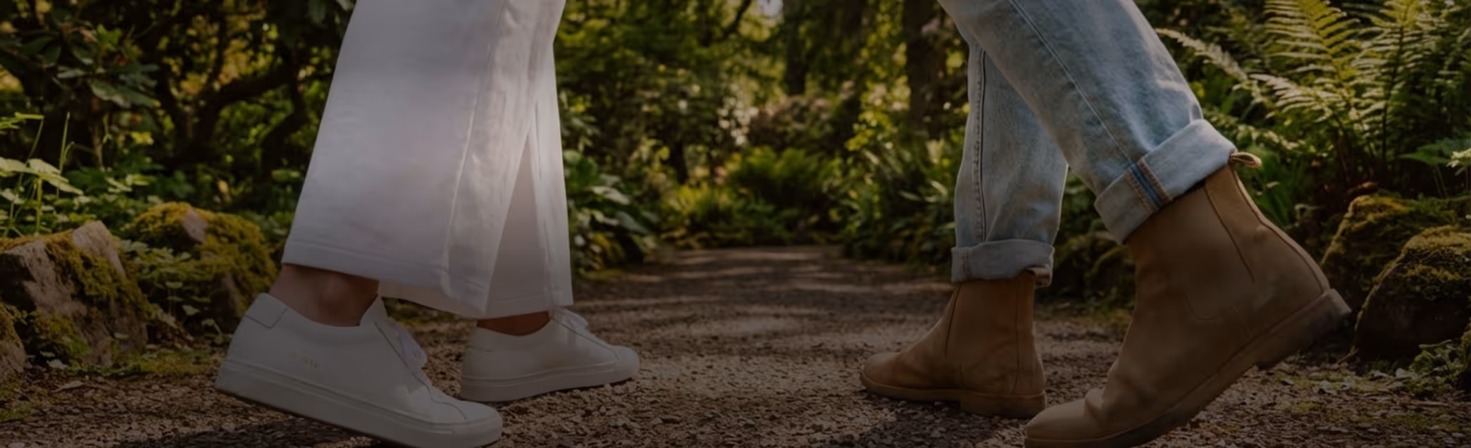 Close-up of two people walking on a gravel path surrounded by green plants, wearing white sneakers and tan boots with cuffed jeans.