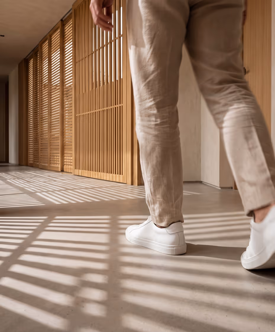 Person wearing beige pants and white sneakers walking on floor with striped shadows from wooden slats.