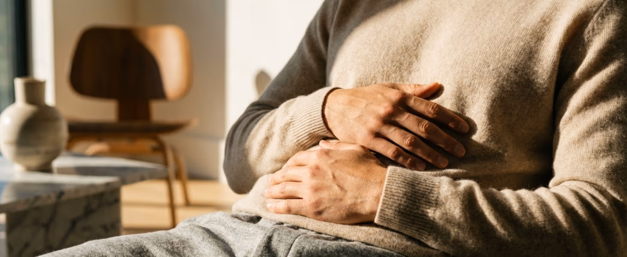 Person sitting indoors holding their stomach, suggesting abdominal discomfort or pain.