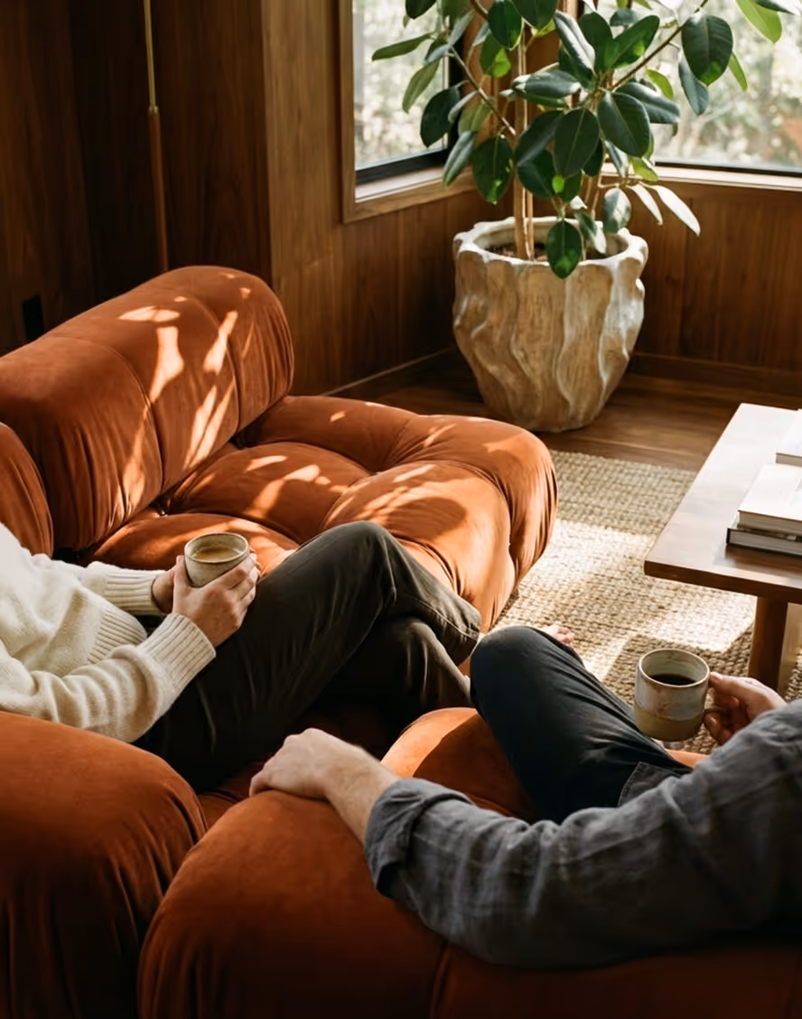 Two people sitting on orange sofas in a sunlit room, each holding a cup of coffee or tea.