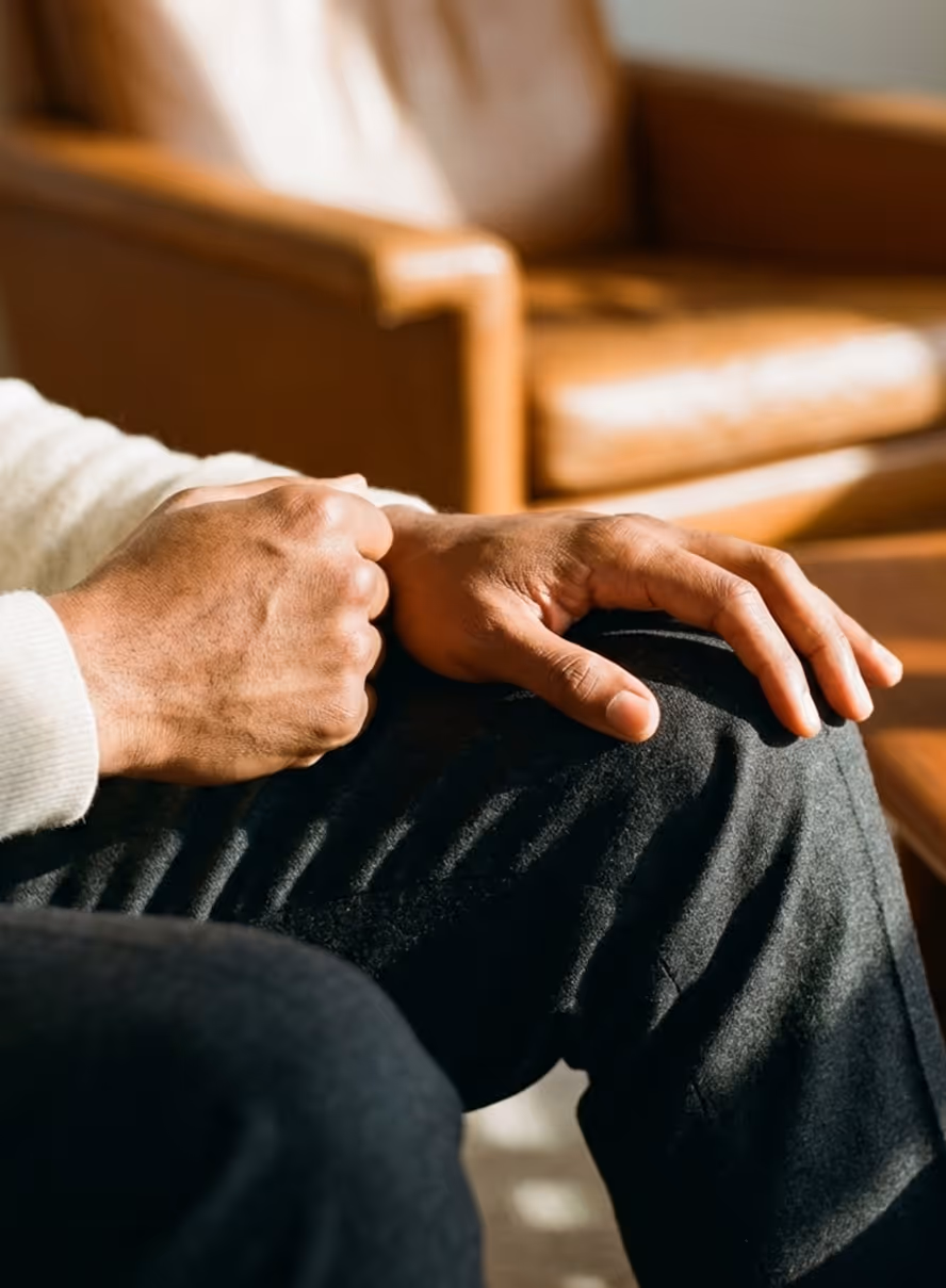 Close-up of a person sitting with hands resting on their knee in a softly lit room.