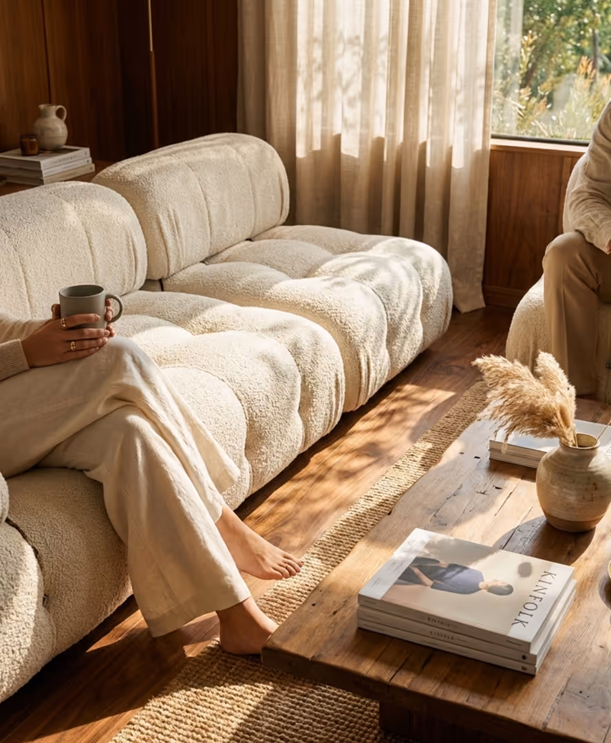 Cozy living room with two beige plush sofas, a wooden coffee table with books and a vase, and sunlight streaming through sheer curtains.