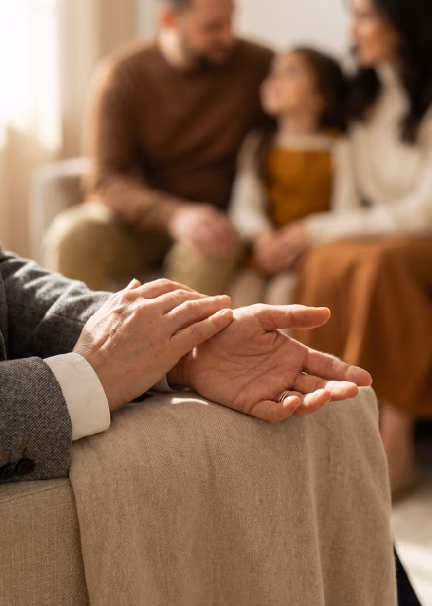 Therapist's hands resting on their knee in the foreground with a family of two adults and a child sitting together in the blurred background.
