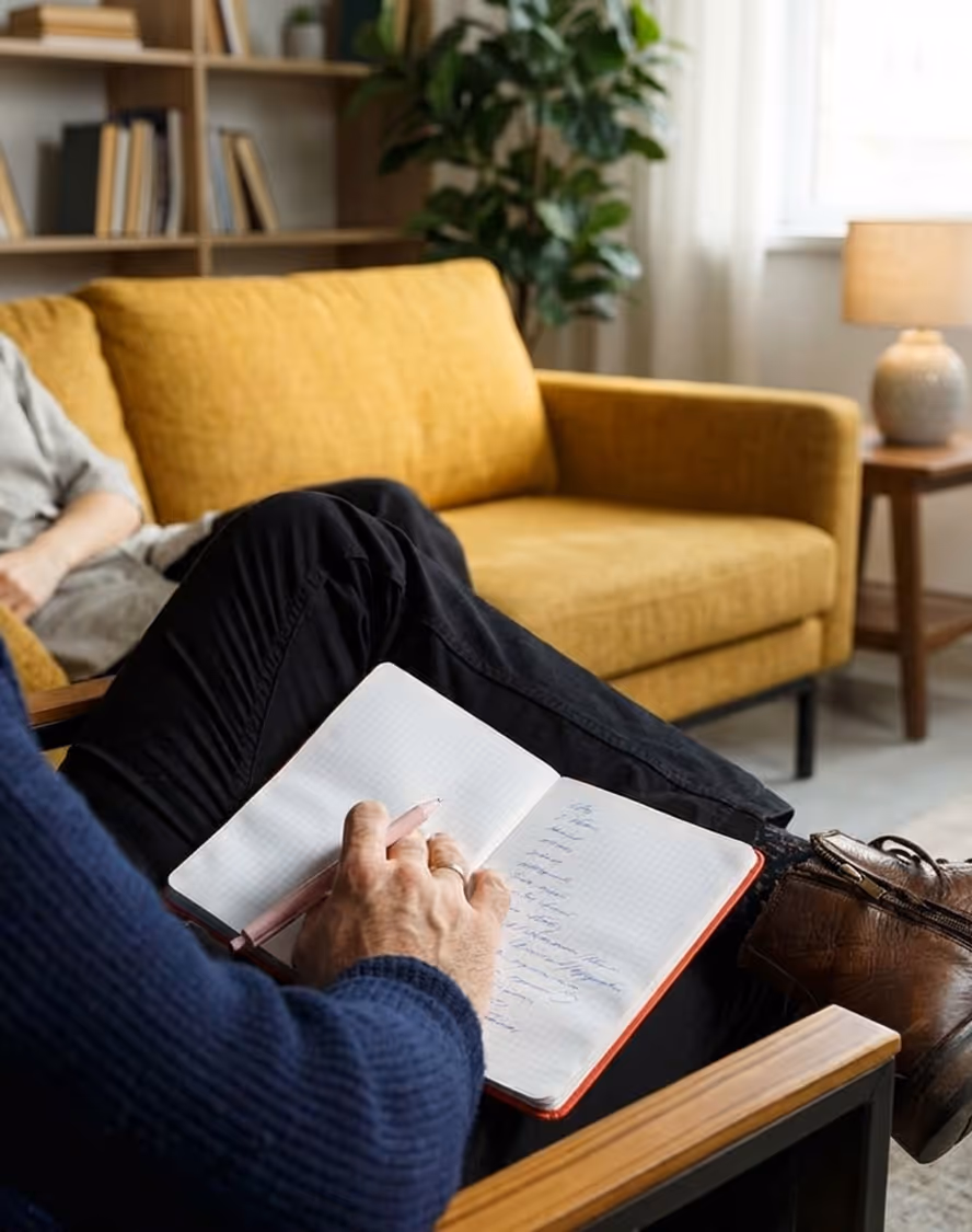Person sitting on a chair writing notes in a notebook with a yellow sofa and bookshelf in the background.