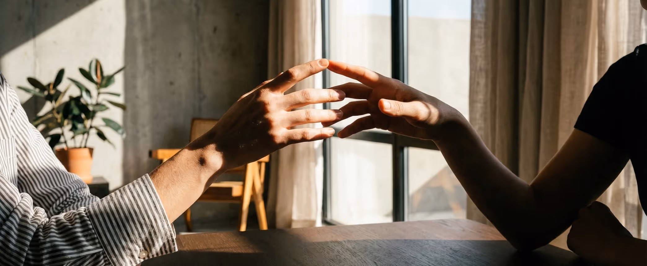 Two people reaching out and touching fingertips across a table near a window with sunlight.