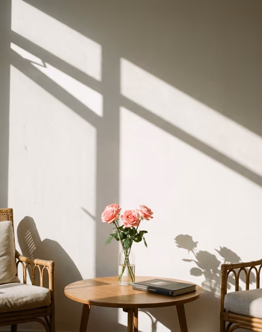Round wooden table with a vase of pink roses and a closed book, flanked by cushioned rattan chairs, with sunlight casting window shadows on a white wall.