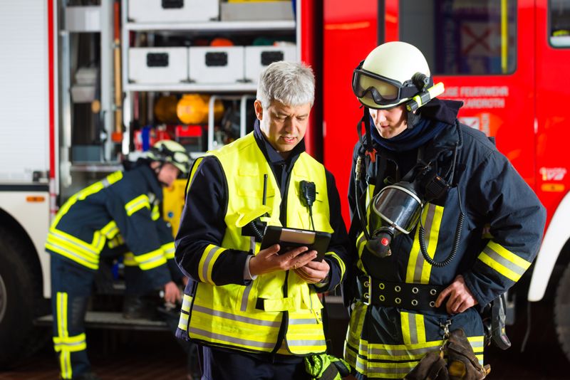 A firefighter in full gear and a colleague in high-visibility vests conferring beside a fire engine