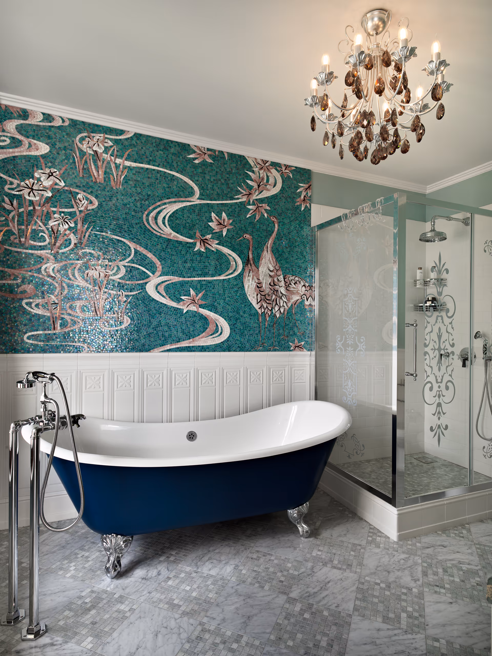 Elegant bathroom with a blue clawfoot bathtub, decorative green tile mural featuring birds, glass shower enclosure with etched patterns, marble floor tiles, and a chandelier.