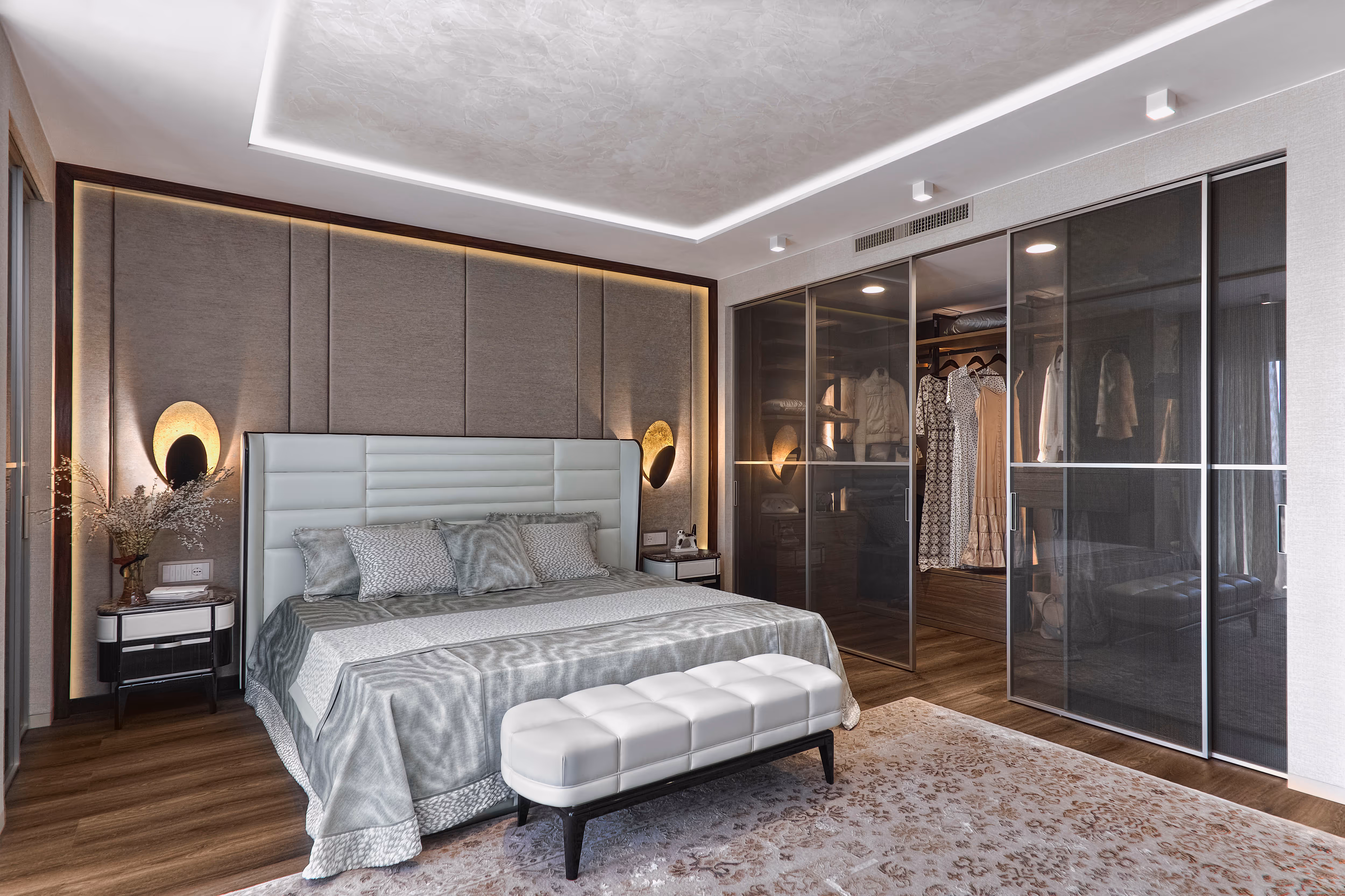 Modern bedroom with a large bed, white leather bench, padded headboard, nightstands with wall lamps, and a glass-door closet showing hanging clothes.
