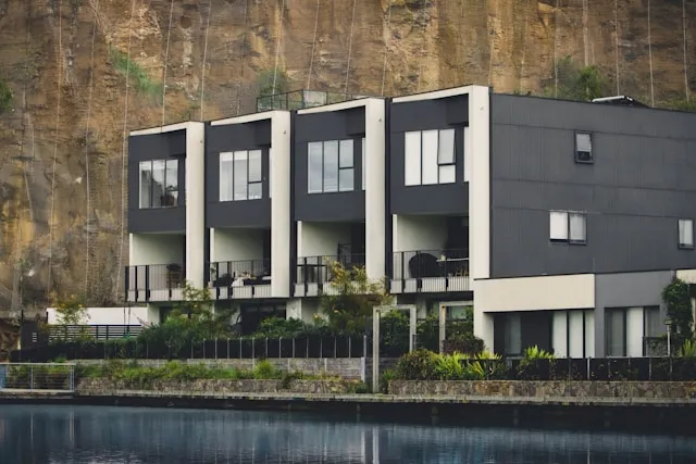 Modern multi-unit residential building with large windows and balconies, set against a steep rocky cliff and overlooking calm water.