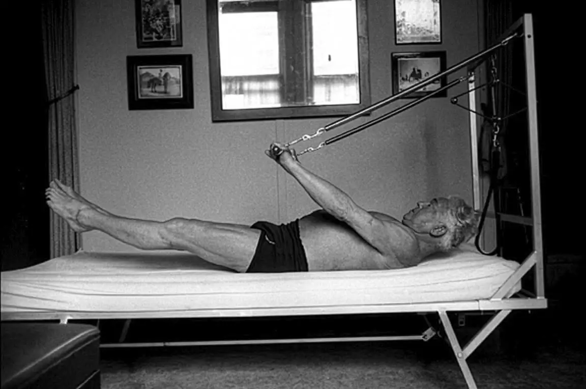 Schwarzweiß-Foto von Joseph Pilates, der auf einem Pilates-Reformer trainiert, mit gestreckten Beinen und Armen, die an Widerstandsbändern ziehen.
