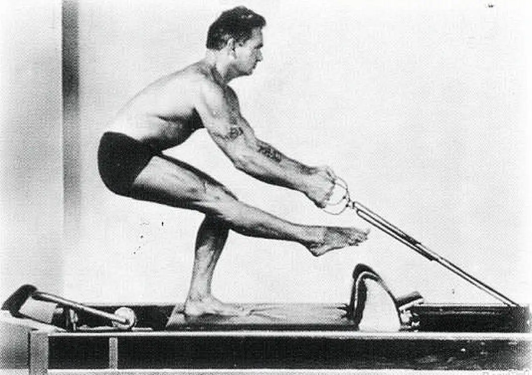 Schwarzweiß-Foto von Joseph Pilates in kurzer Hose, der auf einem Pilates-Gerät steht und mit ausgestrecktem Bein trainiert.