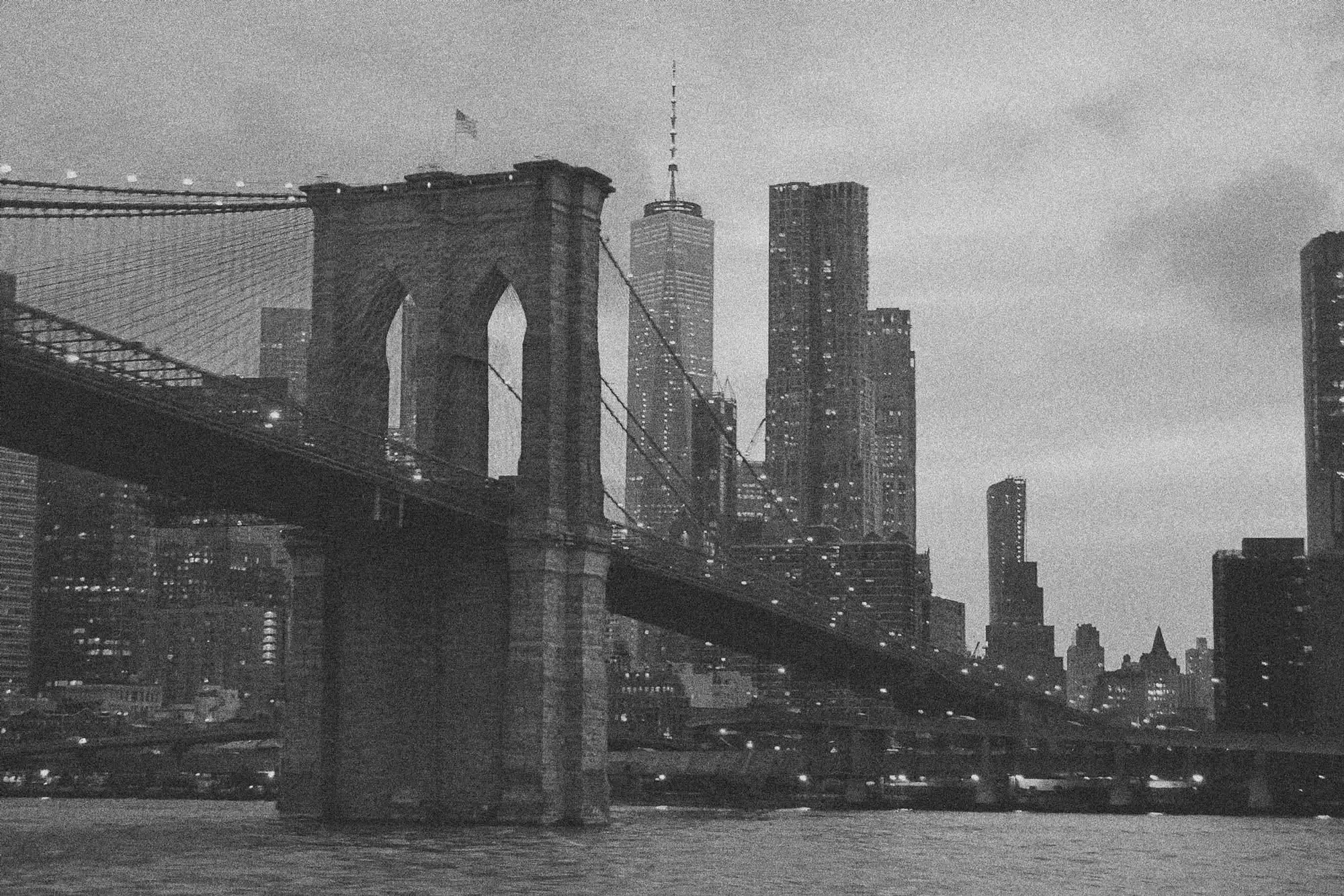 Schwarz-weiß-Aufnahme der Brooklyn Bridge bei Dämmerung mit beleuchteter Stadtsilhouette im Hintergrund.