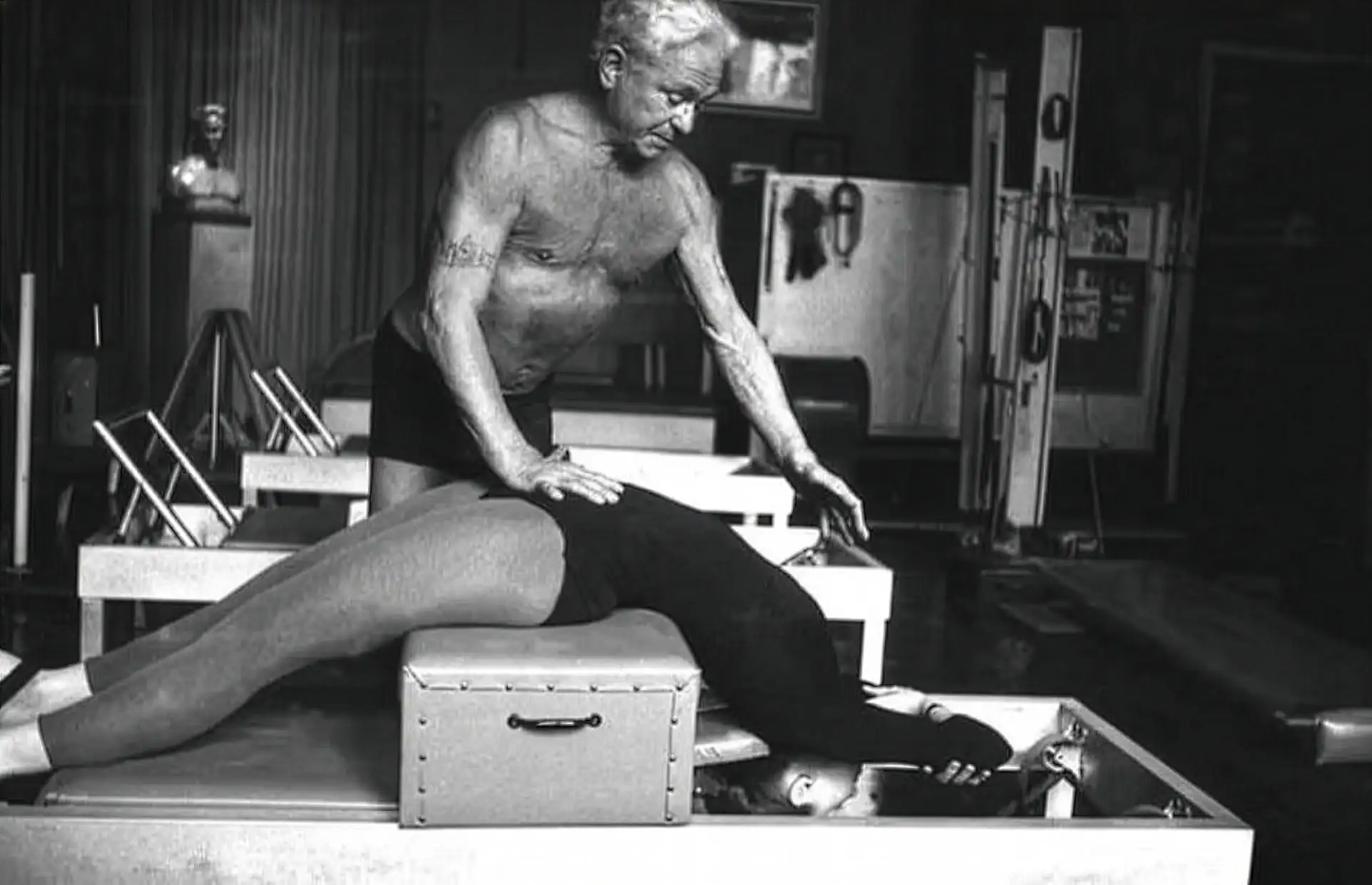 Schwarzweiß-Foto von Joseph Pilates, der einer Frau bei einer Dehnübung auf einem Pilates-Gerät anleitet.