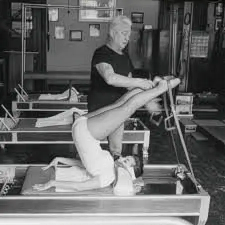 Schwarzweißfoto zeigt eine Frau, die auf einem Pilates-Reformer-Gerät trainiert, während Joseph Pilates ihre Beine hält.