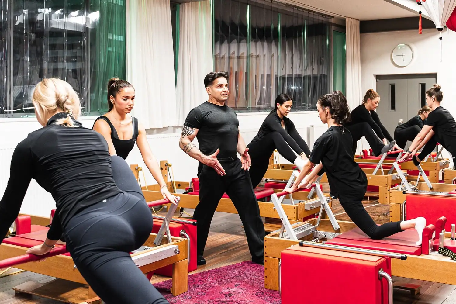 Eine Gruppe von Menschen trainiert Pilates auf Reformer-Geräten in einem hellen Studio unter Anleitung eines Trainers.