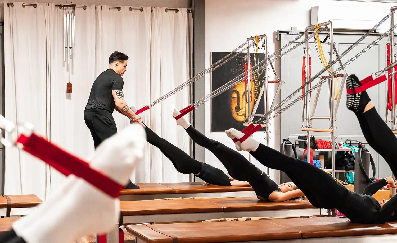 Pilates-Kurs mit Trainer, der Teilnehmern hilft, Beine an Seilen auf dem Pilates-Gerät Wall Unit (Matte) zu strecken.