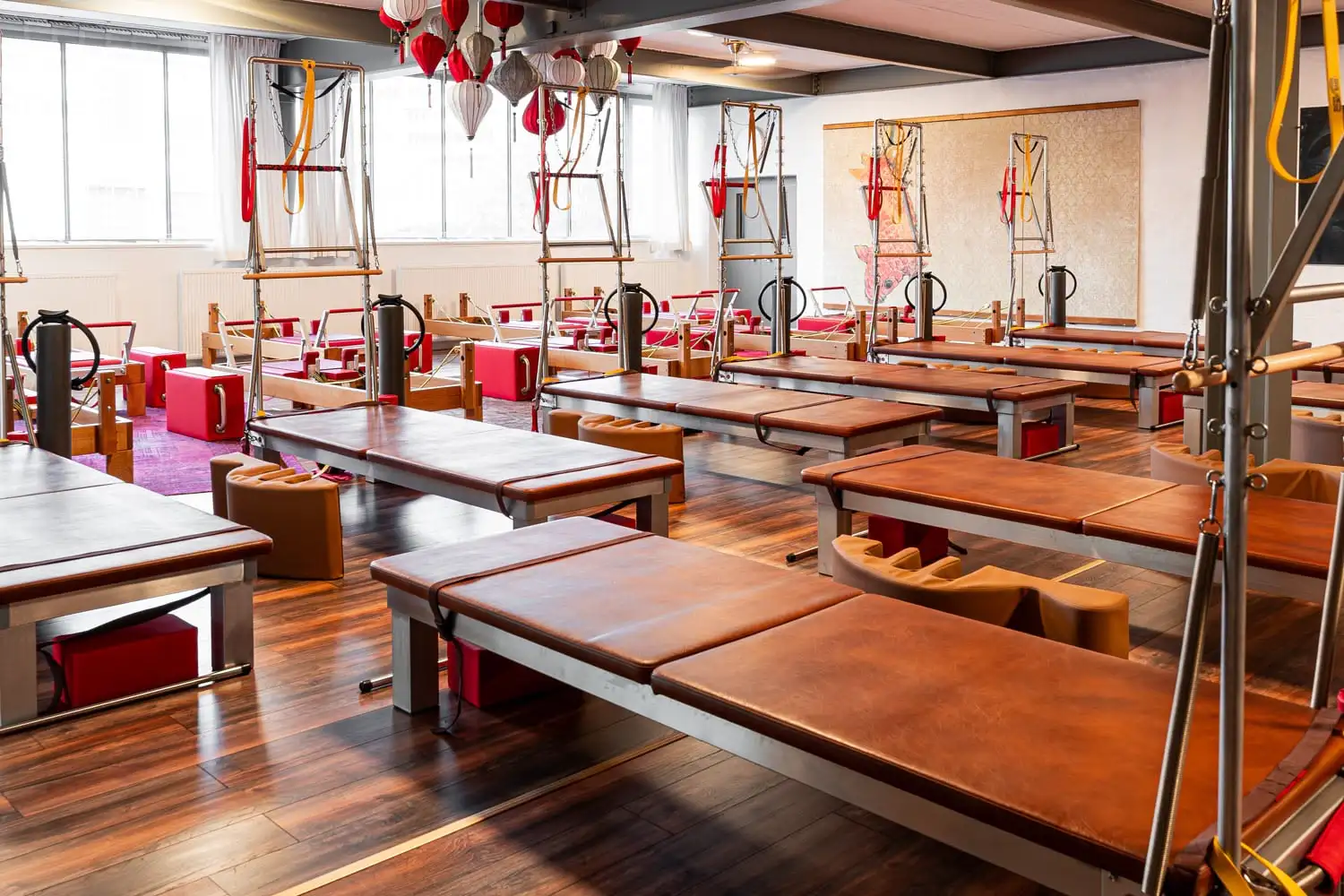 Helles Pilates-Studio in Hannover mit Holzboden und mehreren gepolsterten Reformer-Geräten mit roten Akzenten und Wall Unit (Matte) Geräten.