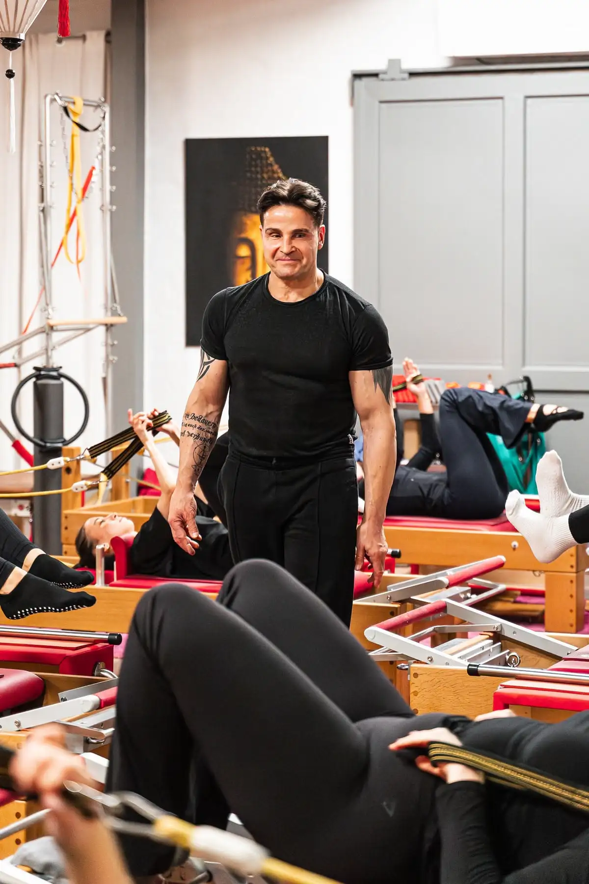 Pilates Trainer in schwarzem T-Shirt, der in einem Pilates-Studio mehrere Personen auf Reformer-Geräten trainiert.