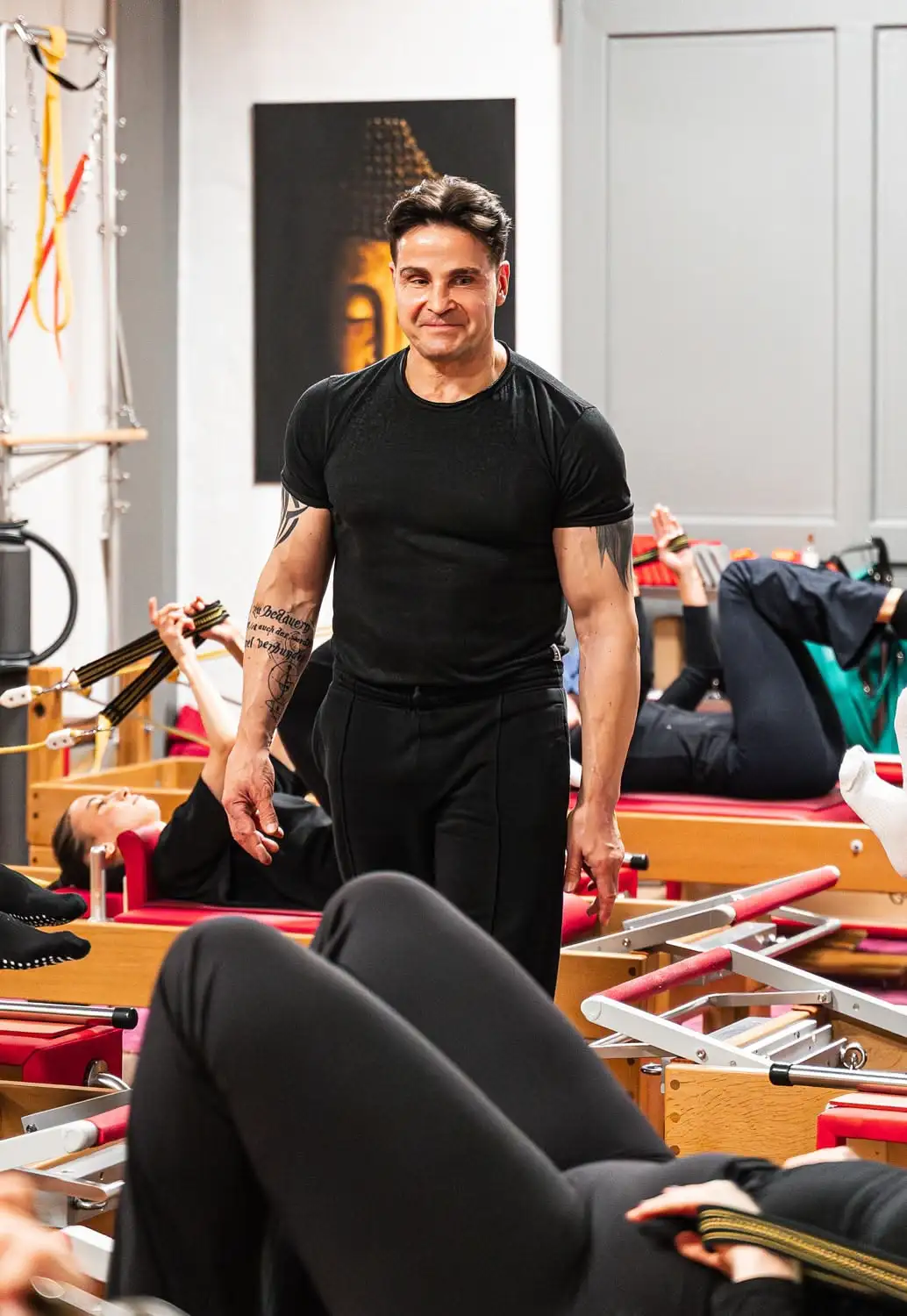 Ein Pilates Trainer im schwarzem T-Shirt beobachtet Teilnehmer während einer Pilates-Übung auf Reformer-Geräten in einem Pilates Studio in Hannover.