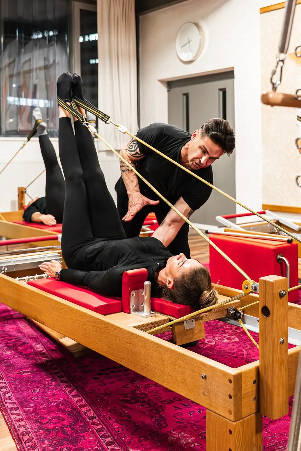 Frau liegt auf einem Pilates-Reformer-Gerät und hebt die Beine, während ein Pilates Trainer Anweisungen gibt.