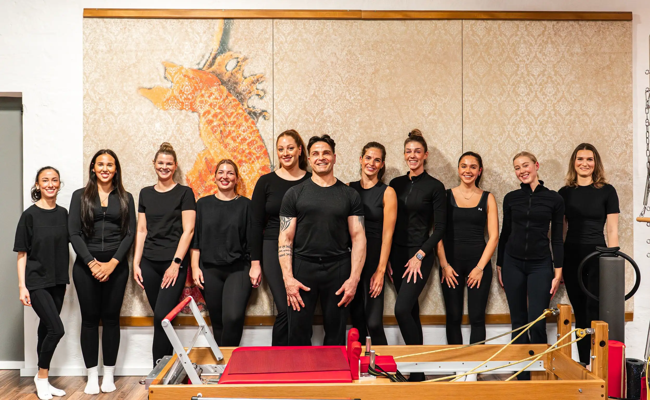 Gruppe von elf Personen in schwarzer Sportkleidung steht in einem Pilates Studio vor einer Wand mit großem orangefarbenem Kunstwerk neben einem Pilates-Reformer-Gerät.