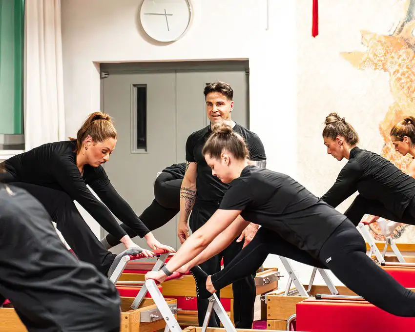Drei Frauen trainieren auf Pilates-Reformern, der Trainer in schwarzem T-Shirt beobachtet und lächelt.