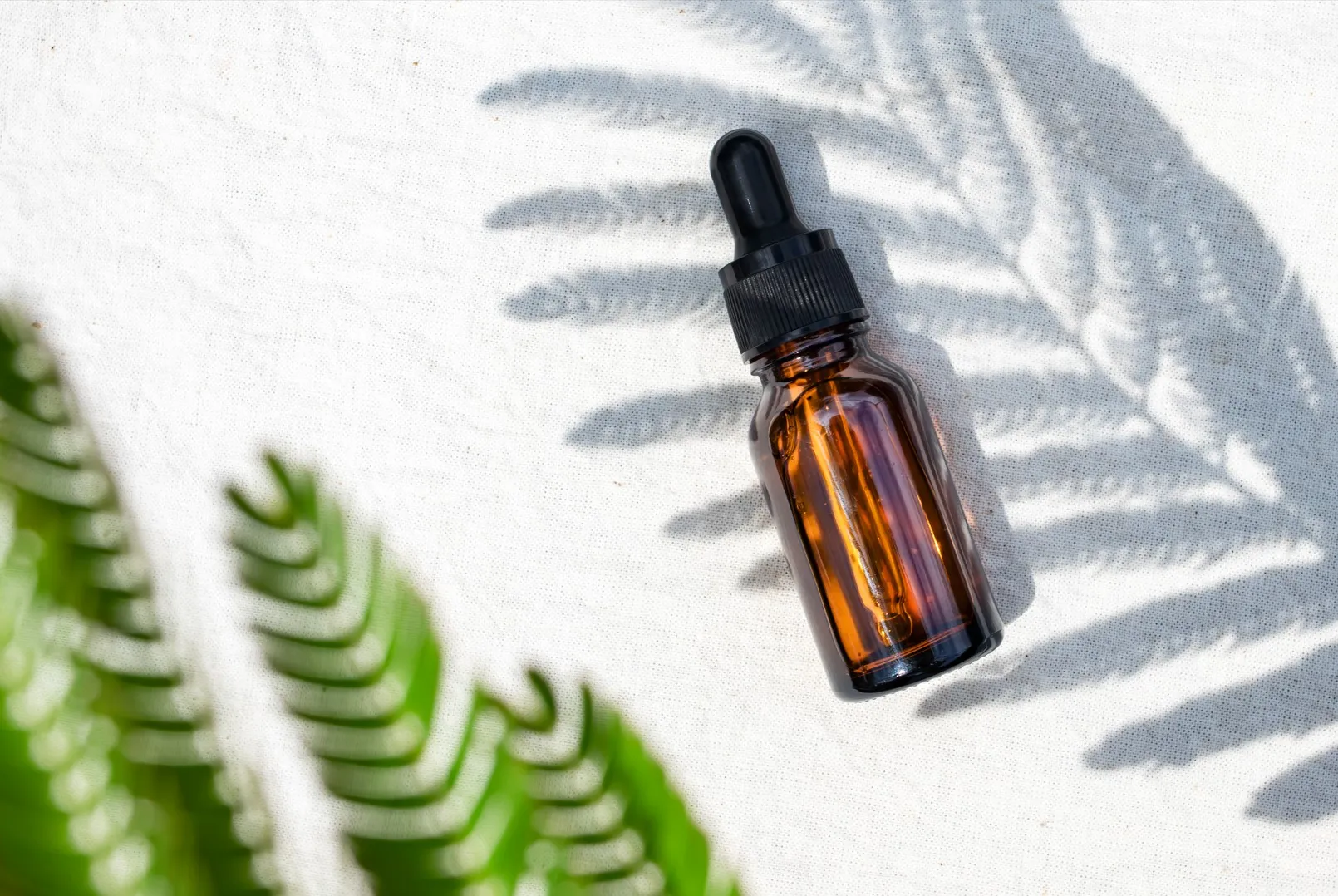 Braune Glasflasche mit Tropfer liegt auf weißem Stoff, im Schatten eines Farnblatts, mit grünem Farn unscharf im Vordergrund.