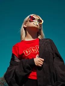 Young woman wearing oversized novelty sunglasses and a red shirt, holding a black jacket against a clear blue sky.